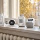 Fünf smarte Heizkörperthermostate von Homematic IP, tado, FRITZ!, Bosch und Eve nebeneinander auf einer Fensterbank über dem Heizkörper im herbstlichen Wohnzimmer